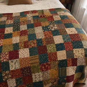 Può includere: Una trapunta patchwork caratterizzata da una griglia di pezzi di tessuto quadrati in vari motivi e colori, tra cui rosso scuro, blu navy, giallo senape, verde oliva e crema. La trapunta mostra una disposizione ripetitiva di stampe floreali.