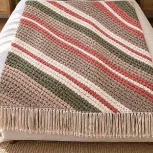 May include: A rectangular crochet blanket featuring diagonal stripes in shades of sage green, dusty rose, cream, and taupe. The textile has a textured stitch pattern and is finished with long, cream-colored fringe along the bottom edge.