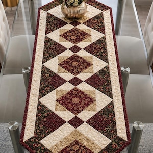 May include: A rectangular quilted table runner featuring a geometric pattern of alternating diamonds and squares. The fabric displays a color palette of deep burgundy, dark forest green, and cream, with intricate floral and paisley-style prints.