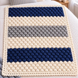 Könnte beinhalten: Eine rechteckige Häkeldecke mit einem strukturierten Noppenmuster, das in horizontalen Streifen in Marineblau, Creme und Grau angeordnet ist. Die Decke ist an allen vier Kanten mit einer dekorativen, cremefarbenen, gewellten Bordüre versehen.