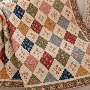 May include: A quilted blanket featuring a diamond pattern of alternating cream-colored squares and squares in shades of navy blue, sage green, dusty rose, and mustard yellow, all decorated with small floral prints and finished with a floral border.