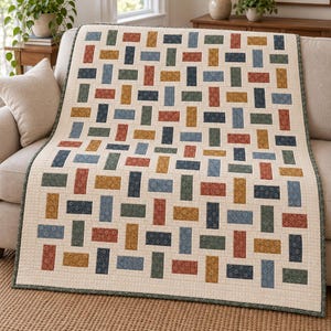 May include: A rectangular quilt featuring a repeating pattern of small, solid-coloured fabric rectangles in shades of rust, mustard yellow, sage green, and navy blue, all set against a cream-coloured background with a subtle grid-like texture.