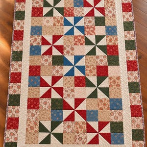 May include: A rectangular quilt featuring a pinwheel pattern in red, green, and blue, interspersed with floral-patterned beige squares. The quilt is framed by a cream-colored border and an outer edge of alternating red and green squares.