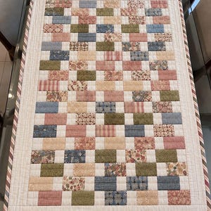 May include: A rectangular quilted table runner featuring a brick-patterned design composed of small fabric rectangles in muted shades of sage green, dusty rose, light blue, and cream. The runner has a solid cream border and a striped edge binding.