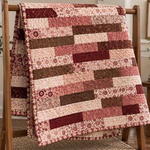 May include: A rectangular quilt featuring a brick-like pattern of alternating fabric strips. The design incorporates various shades of burgundy, dusty rose, and cream, with floral and geometric prints. The entire surface has a quilted texture.