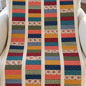 May include: A rectangular quilt featuring four vertical columns of horizontal fabric strips. The strips display a variety of patterns including floral prints, gingham checks, and solid colors in shades of red, blue, green, and yellow, separated by cream-colored sashing.