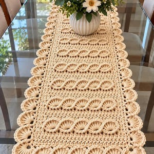 Pode incluir: Um caminho de mesa retangular de crochê na cor creme, com um padrão trançado repetitivo no centro e uma borda festonada em ponto concha nas laterais. Um vaso branco canelado com flores brancas está posicionado sobre o caminho.