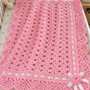 Puede incluir: Una manta rectangular de ganchillo rosa con un intrincado patrón calado, un borde festoneado y una cinta de satén blanca tejida por los bordes. Una flor de ganchillo rosa decorativa con extremos de cinta blanca está unida a una esquina.