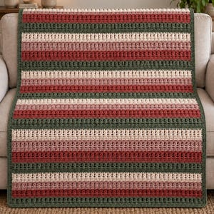 May include: A rectangular crochet blanket featuring horizontal stripes in alternating colors of dark forest green, cream, dusty rose, and deep brick red. The textile displays a repeating textured stitch pattern throughout the entire surface area.
