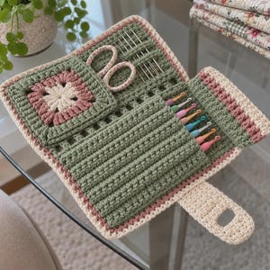 May include: A sage green, dusty rose, and cream crochet organizer case featuring a square motif flap, a pocket for scissors, and multiple slots holding colorful metal crochet hooks and silver needles with a button closure tab.