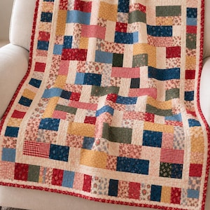 May include: A rectangular quilt featuring a patchwork design of various fabric rectangles in shades of red, blue, yellow, and green, accented with floral and gingham patterns, finished with a red border featuring a small white floral print.