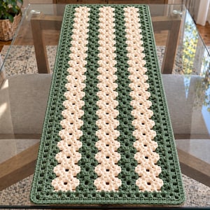 May include: A rectangular crochet table runner featuring alternating vertical stripes of dark forest green and cream-colored yarn. The design consists of a repeating granny square stitch pattern, creating a textured, open-work fabric with a border.