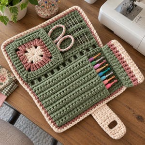 May include: A crochet tool organizer featuring a sage green and cream color scheme with a square motif pocket. It contains a pair of floral scissors, several metal sewing needles, and a row of colorful crochet hooks tucked into integrated slots.