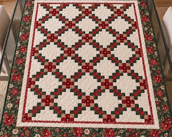 May include: A rectangular quilt featuring a cream-colored center with a repeating grid pattern of red and dark green squares. The border is a dark green fabric decorated with a repeating print of red and white poinsettia flowers.