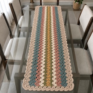 Puede incluir: Un camino de mesa rectangular de ganchillo con un patrón de punto granny repetido y rayas verticales en tonos beige, rosa empolvado, verde salvia y amarillo mostaza, acabado con un borde festoneado en hilo beige claro.