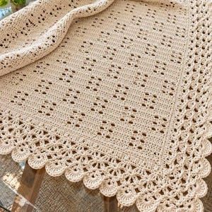 May include: A cream-colored crocheted blanket featuring a repeating square pattern throughout the center. The edges are finished with a decorative scalloped border consisting of rounded, fan-like stitches that create a soft, textured finish.