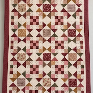 May include: A rectangular quilt featuring a repeating pattern of stars and nine-patch blocks. The design incorporates various fabrics in shades of burgundy, pink, olive green, and mustard yellow, all set against a cream background with a wide burgundy border.