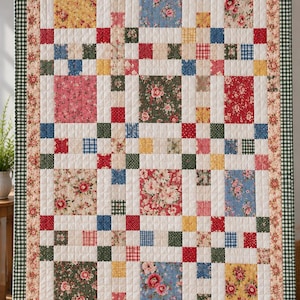 May include: A rectangular patchwork quilt featuring a grid of alternating floral and solid-coloured fabric squares in shades of red, blue, yellow, and green, bordered by a floral print and a dark green and white gingham edge, hanging from clips.