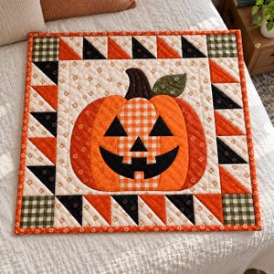 May include: A square quilted wall hanging featuring a central jack-o'-lantern pumpkin with a black face and orange gingham center. The border consists of alternating orange and black triangles with green and white checkered corner squares.