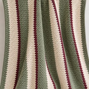 May include: A crocheted blanket featuring vertical stripes in sage green, cream, and deep burgundy. The textile displays a consistent textured stitch pattern throughout, with the stripes arranged in a repeating sequence across the entire surface.