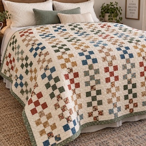 May include: A bed quilt featuring a repeating nine-patch block pattern in a palette of muted red, blue, green, and tan floral prints on a cream background. The quilt is finished with a thin green border and displays a consistent grid design.