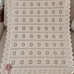 Könnte beinhalten: Eine rechteckige, cremefarbene Häkeldecke mit einem sich wiederholenden Muster aus durchbrochenen Blumenmotiven in horizontalen Reihen. Die Decke ist mit einem dekorativen Bogenrand und einer einzelnen altrosa Stoffblume verziert.