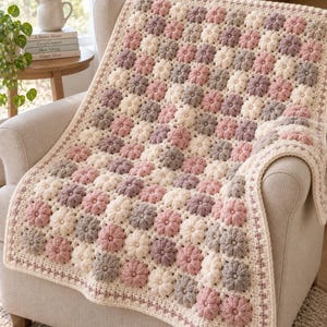 May include: A handmade crochet blanket featuring a repeating pattern of puff-stitch flowers in shades of cream, dusty pink, and muted mauve. The blanket has a textured surface with a thick, cream-coloured border and is draped over a neutral armchair.