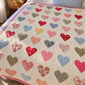 Puede incluir: Una colcha acolchada que presenta una cuadrícula de parches de tela en forma de corazón con diversos estampados florales, de cuadros y lisos. Los corazones están sobre un fondo crema con costuras en forma de diamante y un borde a cuadros rojos y blancos.