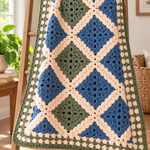 Peut inclure: Une couverture au crochet présentant un motif en losanges composé de carrés alternés vert sauge et bleu roi. Les carrés sont assemblés par une bordure en fil crème, créant un effet de treillis. Les bords sont finis par une bordure vert sauge.