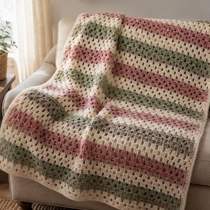 May include: A crochet blanket featuring a granny square stitch pattern with horizontal stripes in alternating colors of cream, dusty rose pink, and sage green. The textile has a textured, open-weave construction with a finished border edge.