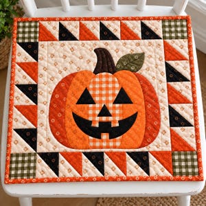 May include: A square quilted wall hanging featuring a central jack-o'-lantern pumpkin with a black face, orange gingham, and floral fabric. The border consists of a geometric pattern of orange, black, and cream triangles and green gingham corners.