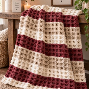 Könnte beinhalten: Eine gehäkelte Decke mit Waffelmuster und abwechselnden horizontalen Streifen in tiefem Burgunderrot, Creme und hellem Beige. Das Textil hat eine dicke, strukturierte Gitteroptik und ist über eine Holzoberfläche drapiert.