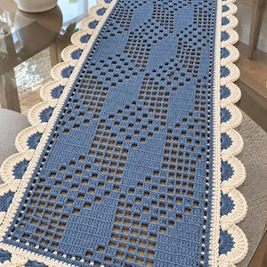 Könnte beinhalten: Ein rechteckiger Häkel-Tischläufer mit einer blauen Mitte in geometrischem Gittermuster und einem cremefarbenen, gewellten Rand. Der Läufer hat ein sich wiederholendes Durchbruchmuster im Mittelteil, das von einer dekorativen Kante aus cremefarbenen Häkelmuscheln eingerahmt wird, die sich mit kleinen blauen Akzenten abwechseln.