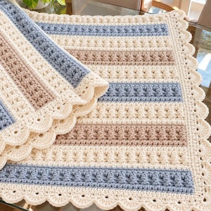 May include: A rectangular crochet blanket featuring alternating horizontal stripes in cream, dusty blue, and light tan. The design incorporates textured bobble stitch patterns and a decorative scalloped edge border in cream yarn throughout.