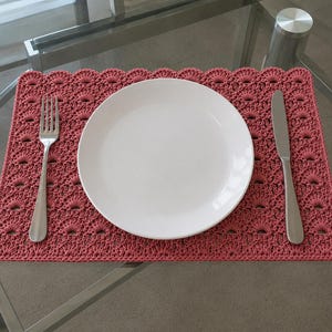 May include: A rectangular, dusty rose crochet placemat featuring a repeating fan-like stitch pattern with a scalloped edge. Centered on the placemat is a plain white round dinner plate, flanked by a stainless steel fork on the left and a knife on the right.