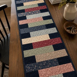 May include: A rectangular quilted table runner featuring a patchwork design of alternating horizontal fabric strips. The pattern includes solid navy blue, muted sage green, dusty pink, and floral-printed fabrics with intricate stitching.