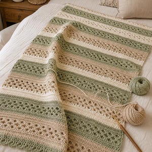 Puede incluir: Una manta de ganchillo rectangular con rayas horizontales alternas en verde salvia, crema y beige. El diseño incorpora filas sólidas y patrones de encaje calado intrincados, terminados con un borde festoneado. Dos ovillos y un ganchillo de madera.