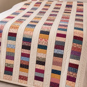 May include: A quilt featuring vertical strips of multicolored fabric patches in various floral, plaid, and solid patterns. The strips are separated by solid cream-colored fabric bands, with visible quilted stitching throughout the entire surface.