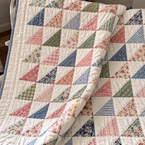 May include: A patchwork quilt featuring a repeating triangle pattern in various floral, striped, and polka dot fabrics. The colour palette includes soft pink, sage green, and dusty blue tones, all set against a solid white background with stitching.