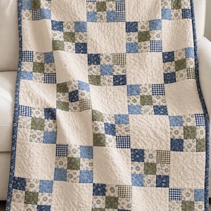 May include: A patchwork quilt featuring a repeating grid pattern of cream-colored squares alternating with blocks of smaller squares in shades of blue, sage green, and white. The quilt has a blue floral border and visible quilted stitching.