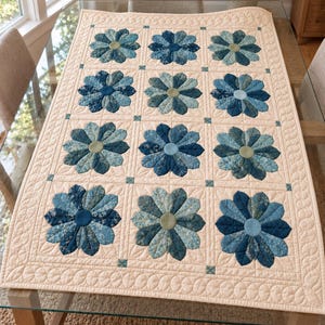 May include: A rectangular quilt featuring a cream-colored background with a grid of twelve flower motifs. Each flower is composed of petals in various shades of blue, ranging from light sky blue to deep navy, with a solid circular center.