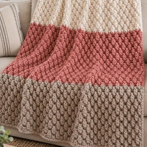 May include: A color-blocked crochet blanket featuring a textured bobble stitch pattern. The blanket is divided into three horizontal sections of cream, dusty rose, and light taupe yarn. The text Crochetspiration.club is visible in the corner.
