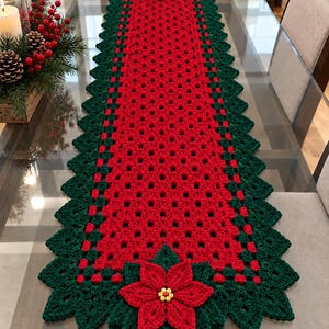 Puede incluir: Un camino de mesa rectangular de ganchillo con un patrón central de rejilla roja bordeado por un borde festoneado de color verde oscuro. El camino está decorado en ambos extremos con una gran flor de pascua roja con centro de cuentas amarillas.