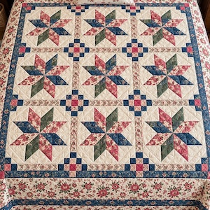 May include: A quilted bedspread featuring a repeating pattern of eight-pointed stars in shades of red, blue, and green on a cream background. The quilt includes floral fabric borders and small square accents, with a matching rectangular throw pillow.