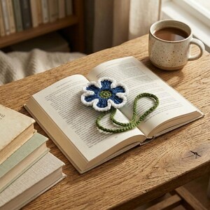 May include: A handmade crochet bookmark featuring a blue flower with a white border and a green center, attached to a long, thin green crocheted stem that rests across the open pages of a book, positioned next to a speckled ceramic mug.