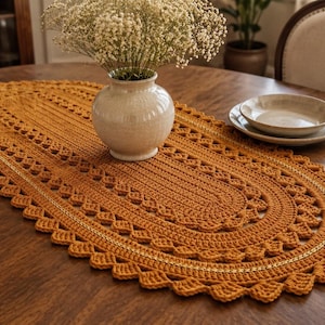 May include: An oval-shaped, mustard-yellow crocheted table runner featuring a textured, concentric stitch pattern with a scalloped edge. The runner is centered on a round wooden table, providing a decorative base for a ceramic vase and dishware.
