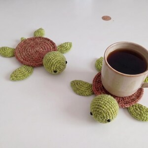 Puede incluir: Dos posavasos de ganchillo hechos a mano con forma de tortuga, con caparazón redondo marrón y cabeza y aletas de color verde claro. Un posavasos sostiene una taza de cerámica beige con café negro, mientras el otro descansa plano cerca.