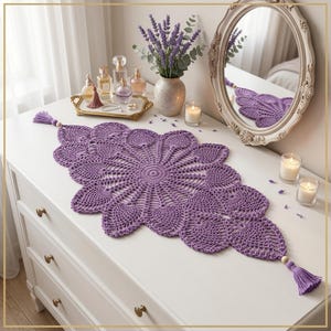 May include: A long, oval-shaped purple crochet table runner featuring an intricate, openwork floral pattern. The runner is finished with a small wooden bead and a matching purple tassel at each pointed end, creating a decorative textile accent.