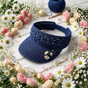 May include: A dark blue crochet visor featuring a decorative band of star-shaped cutouts and a small cluster of white daisy appliques on the brim. The visor is constructed with a dense, textured stitch pattern and an adjustable back strap.