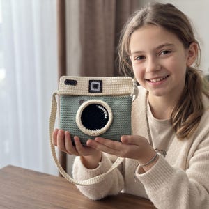 May include: A crochet camera bag featuring a sage green body and a cream-coloured top flap. The bag includes a circular black lens detail with a white border, a cream-coloured shoulder strap, and small rectangular black accents on the top section.